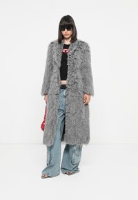 Cappotto in pelliccia sintetica grigia con texture shaggy, design lungo, aperture frontale, indossato sopra un crop top nero e jeans larghi, accessoriato con occhiali da sole.