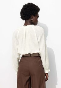 Blouse blanche raccourcie en tissu texturé, à manches longues et avec un col volanté, associée à un pantalon taille haute marron avec des poches arrière.