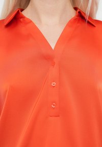 Κόκκινο πουκάμισο με κουμπιά, V-λαιμόκοψη και κοντό γιακά. Διαθέτει πέντε κουμπιά στο ίδιο χρώμα και έχει λεία, γυαλιστερή υφή.