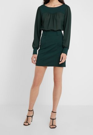 Mujer vestida con una blusa de manga larga verde oscuro y una falda ajustada a juego, con sandalias negras de tacón alto, de pie frente a un fondo liso.