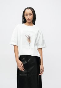 Biała oversize koszulka z rozmytym nadrukiem tancerza na przodzie. W zestawieniu z czarną spódnicą z imitacji skóry o gładkiej fakturze.