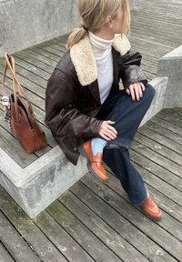 Ruskea nahkatakki, jossa on karvakaulus, kerma värinen pooloneule, tummansiniset leveälahkeiset farkut, meripihkansävyiset loaferit ja ruskea käsilaukku, jossa ruudullinen yksityiskohta.