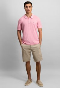 Lyserød poloskjorte med klassisk krave, parret med beige shorts. Modellen står med hænderne i lommerne, iført lysebrune slip-on sko.