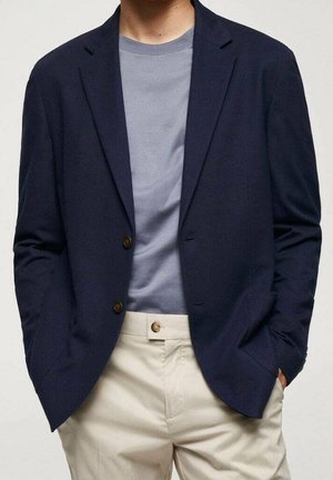 Mand iført en navy blazer over en grå t-shirt og beige bukser med hænderne i lommerne mod en ensfarvet baggrund.