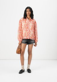 Floral patterned long-sleeve shirt in shades of orange and pink, paired with black denim shorts and black loafers, holding a small brown bag.