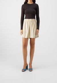 Haut à manches longues, marron foncé, associé à une jupe plissée à taille élastique crème et des sandales à talons bleues, sur un fond neutre.