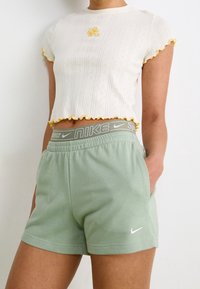 Bílý zkrácený top se žlutými vlnkovanými okraji a květinovou výšivkou, spárovaný se světlými zelenými šortkami Nike s elastickým pasem.