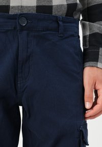 Tmavě modré bavlněné cargo kalhoty s uzávěrem na knoflík, poutky na opasek a bočními kapsami. Tkanina má hladkou strukturu, zobrazeno od pasu dolů.