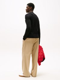 Haut noir à manches longues, pantalon beige à jambes larges, baskets blanches, tenant une veste matelassée rouge. Tissu lisse, design simple, coupe décontractée.