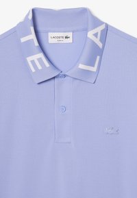 Licht lila polo shirt met een geribbelde kraag en witte letters. Inclusief een placket met twee knopen en een geborduurd logo op de borst.