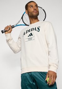 Sweatshirt crème avec un logo "ADIDAS" vert foncé et le texte "TENNIS CLUB". Le modèle tient une raquette de tennis et une balle de tennis jaune, portant un short teal.