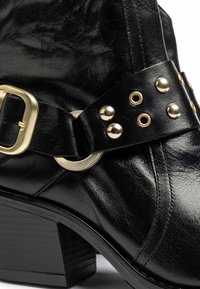 Stivale alla caviglia in pelle nera con superficie texture, decorazioni in metallo dorato, una fascia ampia con borchie e un tacco robusto.