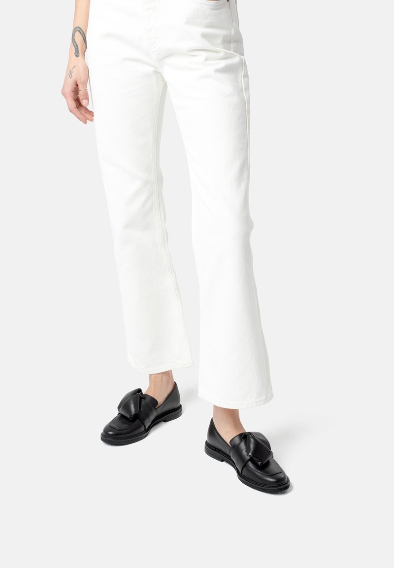 Schwarze Lederloafer mit einer großen Schleifenakzent, kombiniert mit weißen, ausgestellten Jeans. Sie zeichnen sich durch eine glatte Textur und ein rundes Zedesign aus.