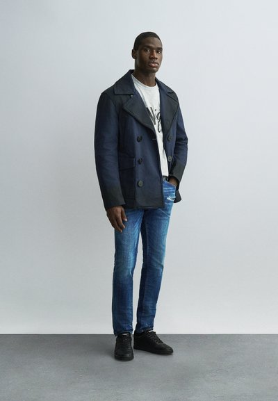 Homme portant un manteau croisé bleu marine, un t-shirt gris clair imprimé, un jean bleu et des baskets noires, debout contre un mur uni gris clair.