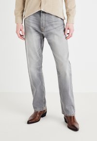Light gray jeans in a relaxed fit with subtle fading details, paired with brown pointed-toe boots, worn with a tan, corduroy shirt.