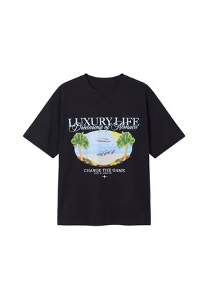 Homme debout portant un t-shirt noir avec des palmiers et un jean large bleu clair sur un fond blanc uni.