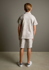 Hellbeiges Kurzarmhemd und passende Shorts mit weißen Blattmustern. Getragen mit weißen Turnschuhen, von hinten gesehen.