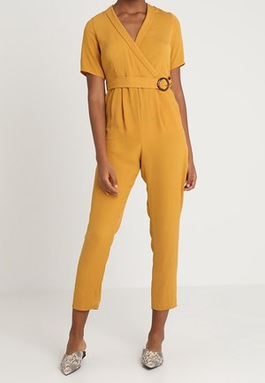 Mujer vistiendo un mono de manga corta color mostaza, con escote en V, cintura con cinturón con hebilla redonda y pantalones pitillo acabados en tobillo.
