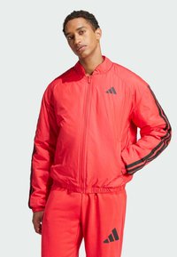 Red insulated bomber jacket with black stripes, zipper front, raised collar, and elastic cuffs. Worn with matching red pants featuring a logo.