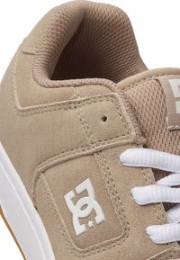 Baskets en daim beige avec lacets blancs, présentant une languette en maille texturée et un patch logo blanc. La semelle en caoutchouc et les détails cousus sont visibles.