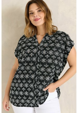 Frau mit schulterlangem Haar, die lächelt, und eine schwarze Kurzarmbluse mit weißer Blumenstickerei sowie weiße Hosen trägt.