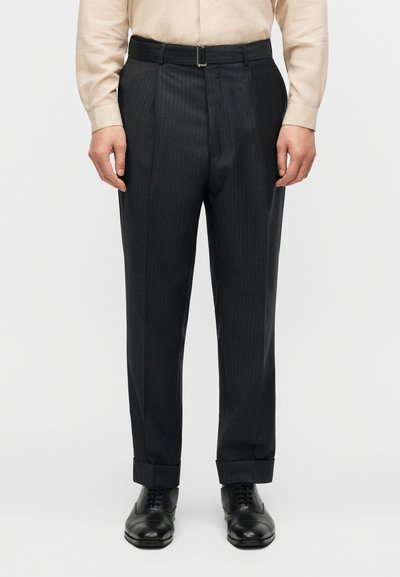 Homme portant un pantalon de costume noir à fines rayures avec ourlets retroussés, chaussures de ville noires, chemise beige à manches longues rentrée, debout devant un fond blanc.