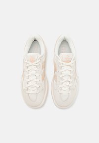 Un par de zapatillas color crema con acentos en melocotón claro, texturas de malla y gamuza, y cordones blancos vistas desde arriba sobre un fondo blanco.
