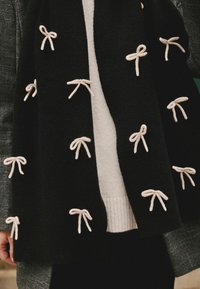 Sort tørklæde med broderede cremede sløjfer med jævne mellemrum, lavet af blødt stof med en tekstureret overflade, båret over en hvid sweater.