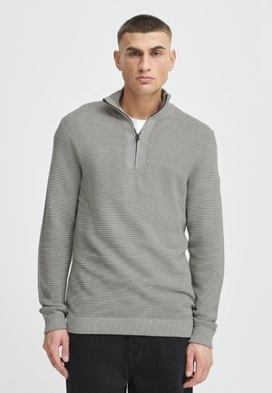 Bărbat cu păr scurt, purtând un pulover gri deschis texturat cu fermoar pe jumătate și pantaloni negri, stând pe un fundal simplu și deschis la culoare.