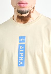 Beige t-paita, jossa on suorakulmainen sininen painatus, jossa lukee "ALPHA" ja sen yläpuolella valkoinen logo. Pyöreä pääntie, lyhyet hihat.