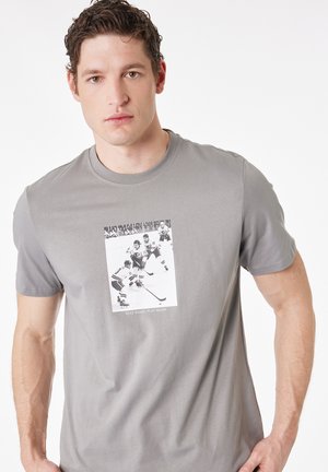 Uomo che indossa una maglietta grigia con foto in bianco e nero di giocatori di hockey e la scritta "STAY SHARP, PLAY SHARP" sul petto, che guarda avanti.