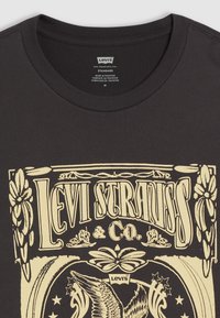 Tricou negru din bumbac cu text grafic de culoare crem, decorat cu modele ornate, inclusiv flori și o vultur, centrat pe partea din față.
