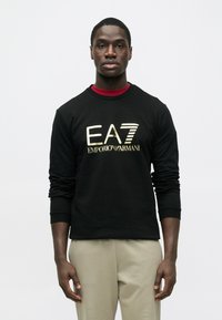 Čierna mikina s dlhým rukávom so zlatým logom "EA7 EMPORIO ARMANI" na prednej strane, v kombinácii so svetlo béžovými nohavicami.