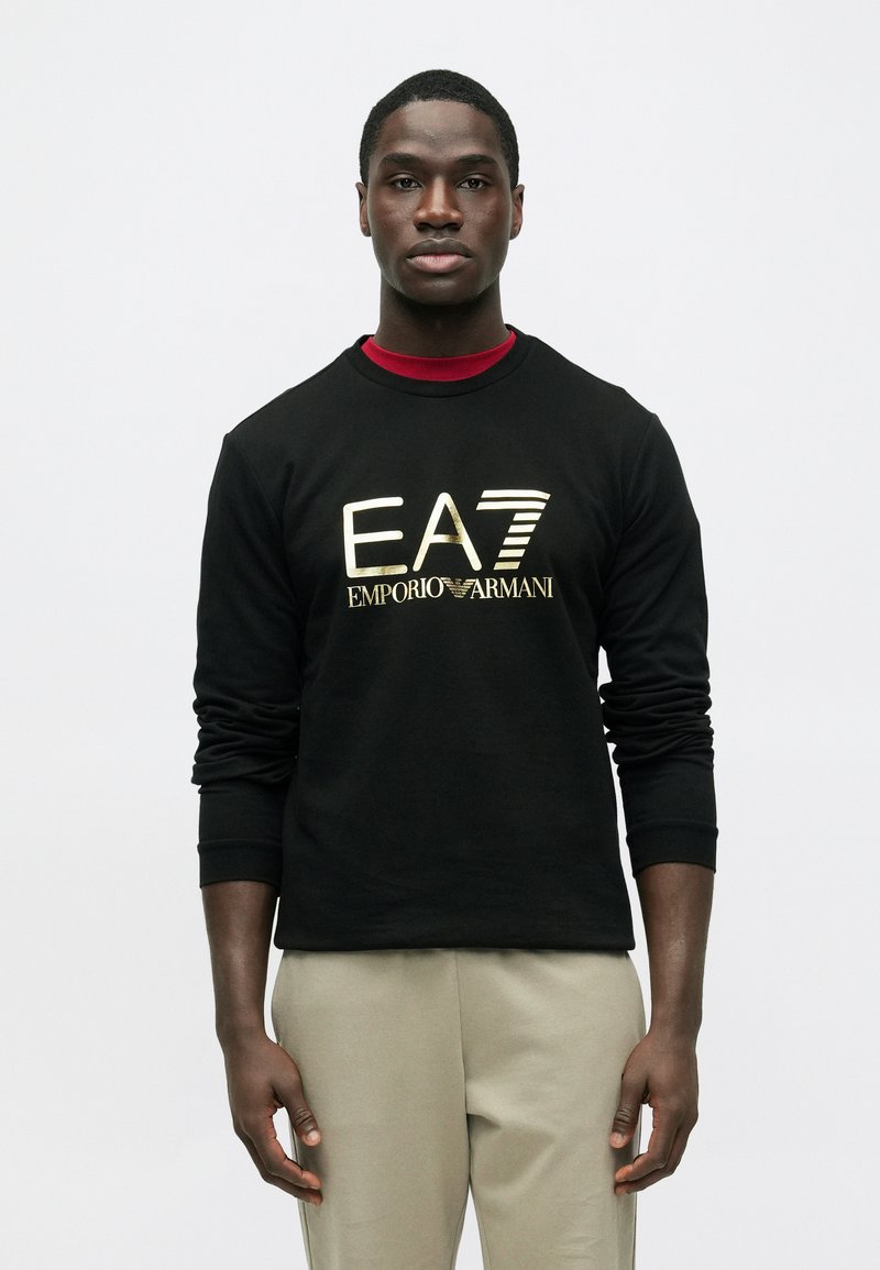 Čierna mikina s dlhým rukávom so zlatým logom "EA7 EMPORIO ARMANI" na prednej strane, v kombinácii so svetlo béžovými nohavicami.