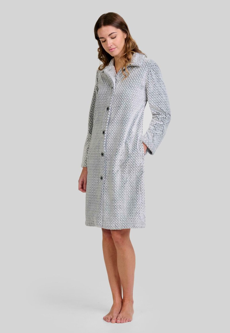 Mujer de pie descalza, vestida con un abrigo texturizado de color gris claro, hasta las rodillas, abotonado, con cuello y bolsillos, sobre un fondo liso gris claro.