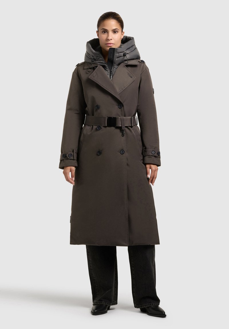 Kvinna som bär en lång mörkbrun bälte trenchcoat över en kolgrå pufferjacka, tillsammans med svarta vida jeans och svarta stövlar.