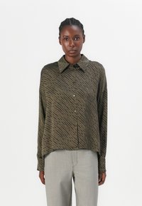Olivgrünes Hemd mit Knopfleiste und einem abstrakten schwarzen Muster, im Oversized-Schnitt, mit langen Ärmeln und klassischem Kragen. Kombiniert mit hellgrauen Hosen.