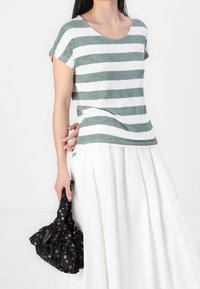 Pruhované zeleno-bílé tričko s krátkým rukávem, zkombinované s volnou bílou sukní. V ruce drží černou květinovou látkovou tašku.