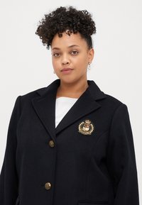 Tengerészkék gyapjúkeverék blézer, strukturált gallérral, aranyszínű gombokkal és díszes címerrel a mellkason. Simább textúrájú és szabott fazonú.