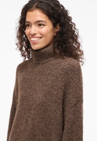 Pull à col roulé marron en tissu tricoté texturé avec une coupe décontractée et un col côtelé, présentant de subtiles variations de couleur.