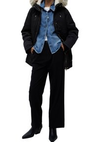 Giacca a vento nera con cappuccio foderato in pelliccia, indossata sopra una camicia in denim azzurro chiaro e una maglietta bianca, abbinata a pantaloni neri a gamba larga e stivali neri con tacco.