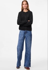 Cardigan preto com botões, de gola redonda, combinado com jeans de perna larga em azul claro e sapatos bordeaux. Textura e detalhes de design mínimos.