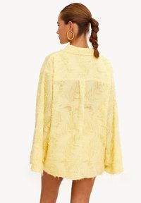 Chemise veste jaune texturée à coupe ample avec manches longues. Elle présente des détails effilochés et un col, avec un dos droit et une poche.