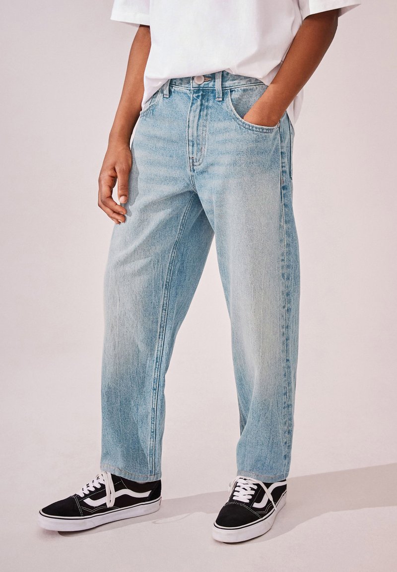 Hellblaue Denim-Jeans mit entspannter Passform, ausgestattet mit zwei Fronttaschen und klassischem Knopfverschluss, kombiniert mit schwarzen Turnschuhen.