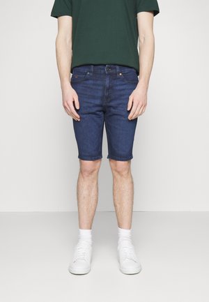 Hombre con pantalones cortos vaqueros de color azul oscuro, calcetines blancos, zapatillas blancas y una camiseta verde oscuro sobre un fondo claro y liso.