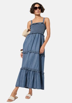 Denim maksi obleka z nabrano spodnjim delom, nabranim korzetom, nastavljivimi naramnicami in zračnim silhuetom. V kombinaciji z natikači in tkanim nahrbtnikom.