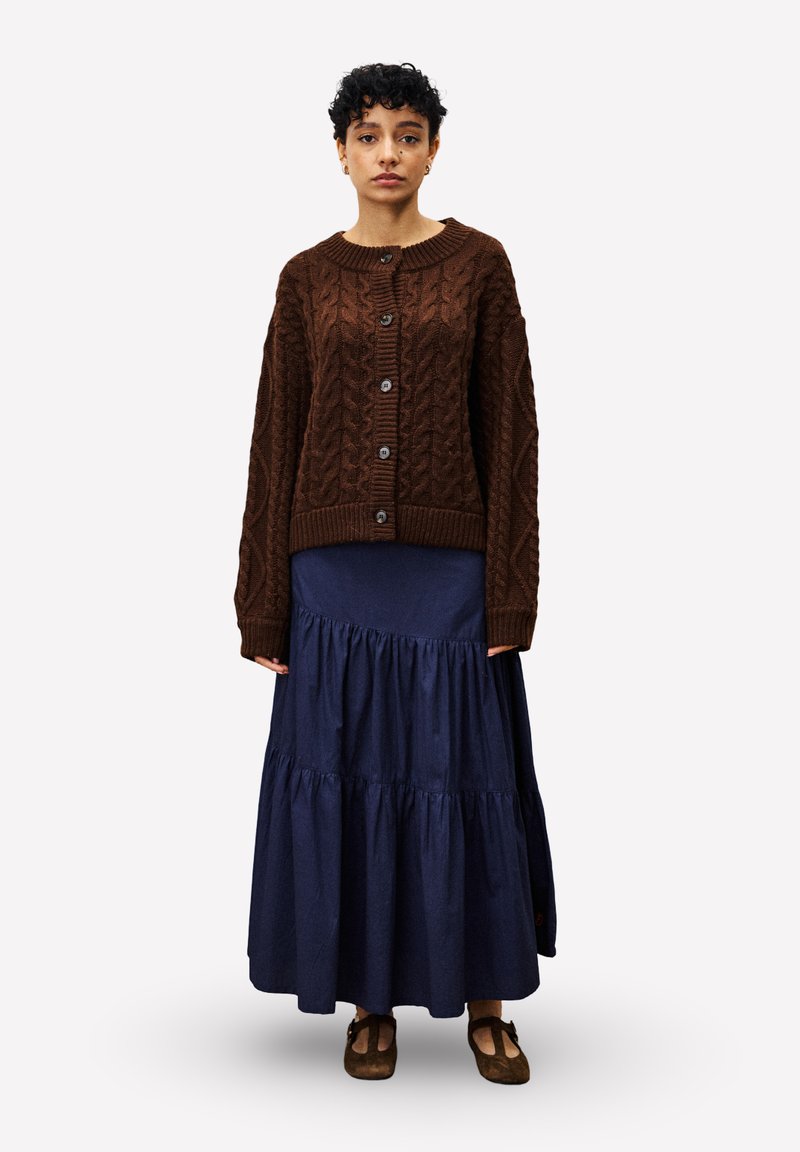 Frau mit kurzen lockigen Haaren, die eine braune Strickjacke mit Zopfmuster, einen langen marineblauen Stufenrock und braune Mary Jane-Schuhe trägt, stehend in Frontalansicht.