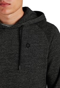 Huvtröja i mörkgrått, tillverkad av strukturert tyg. Har en dragsko i huvan och en liten svart sexkantig logotyp på bröstet.