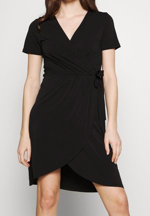 Vrouw draagt een zwart wikkeljurk met korte mouwen, een zijstriksluiting, een V-hals en een asymmetrische zoom.