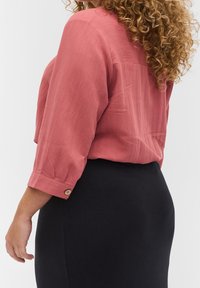 Blouse corail à manches courtes avec une finition texturée et des poignets boutonnés, associée à une jupe noire ajustée. Cheveux bouclés visibles.